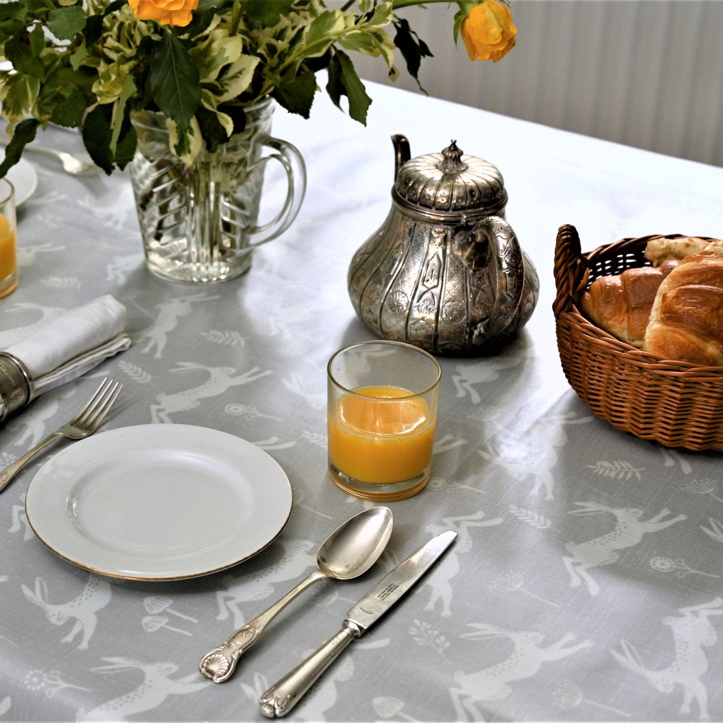A set table featuring a gray tablecloth with a running hare pattern, accompanied by plates, cutlery, glasses of juice, and a basket of bread.